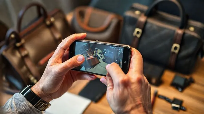 A photorealistic image of a person's hands holding up a used but well-maintained smartphone, in the act of taking a photo for an online selling platform. Other items like a designer bag, a watch, and some electronics are blurred in the background on a table. Cinematic lighting, sharp focus on the phone, 8K hyper-detailed.